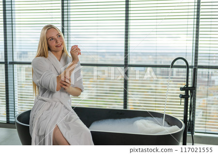Woman applying moisturizing body lotion on hand arm relaxing in bathroom. Girl rubbing nourishing cream doing daily skincare routine spending morning in bathroom. Beauty treatment spa body skin care 117021555