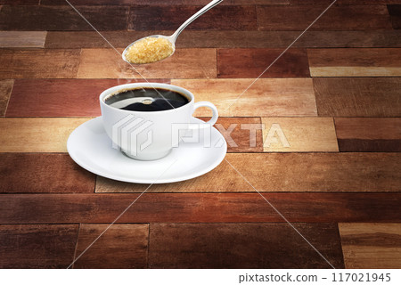 Spoon of sugar poured into a coffee mug with a white saucer on a wooden background. 117021945