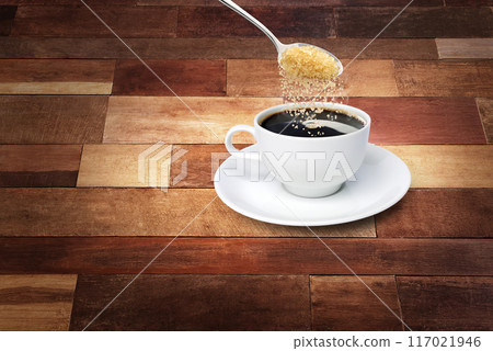 Spoon of sugar poured into a coffee mug with a white saucer on a wooden background. 117021946
