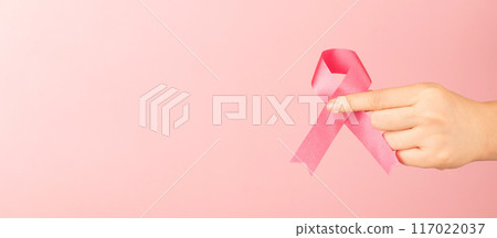 International symbol of Breast Cancer Awareness Month in October. Close up of female hand holding satin pink ribbon awareness on pink background. Women's health care and medical concept. International symbol of Breast Cancer Awareness Month in October. Close up of female hand holding satin pink ribbon awareness on pink background. Women's health care and medical concept. 117022037