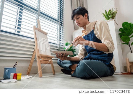 Young Man Painting on Canvas at Home in a Bright Room with Natural Light and Indoor Plants Young Man Painting on Canvas at Home in a Bright Room with Natural Light and Indoor Plants 117023478