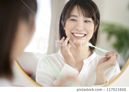 A woman in her early 30s brushing her teeth while looking in the mirror 117023808