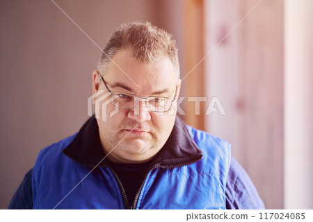 Frowning look from under eyebrows, large chubby-cheeked Caucasian man with glasses looks angrily. 117024085
