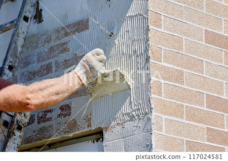 Using steel notched trowel to apply adhesive cement mortar to exterior wall of building before facing faux brick panels. 117024581