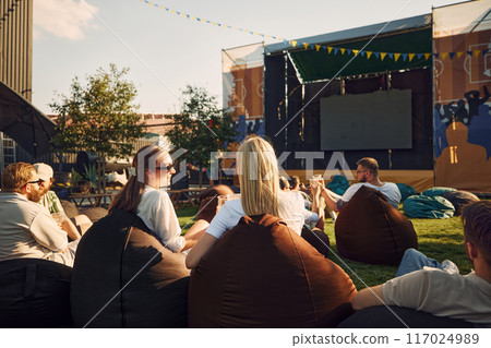 Festival. Group of people, men and women, friends gathering outdoor to watch movie and drink beer on warm summer day 117024989