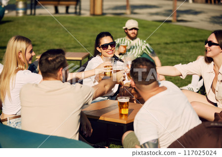 Joyful gathering. Group of friends, men and women sitting outdoors in restaurant, talking, laughing and drinking refreshing beer Joyful gathering. Group of friends, men and women sitting outdoors in restaurant, talking, laughing and drinking refreshing beer 117025024