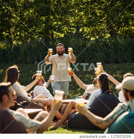 Group of happy smiling young people, friends gathering on outdoor picnic, drinking refreshing drink, having good company 117025030