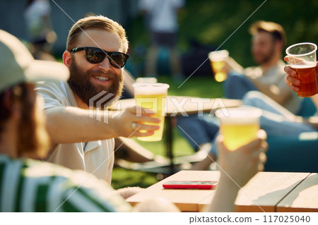 Sunny Celebration. Group of friends, gathering for a picnic, sharing laugh and engaging conversation while clinking beer glasses. Sunny Celebration. Group of friends, gathering for a picnic, sharing laugh and engaging conversation while clinking beer glasses. 117025040