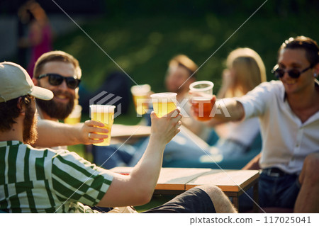 Sunny Celebration. Group of friends, gathering for a picnic, sharing laugh and engaging conversation while clinking beer glasses. Sunny Celebration. Group of friends, gathering for a picnic, sharing laugh and engaging conversation while clinking beer glasses. 117025041
