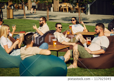 Cheerful young people, friends, men and women gathering on outdoor cafe space, for picnic, enjoying beer and conversations Cheerful young people, friends, men and women gathering on outdoor cafe space, for picnic, enjoying beer and conversations 117025045