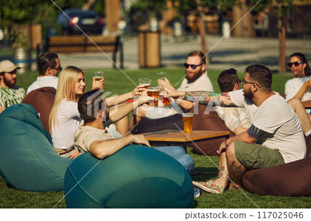 Outdoor picnic in public place. Young people, men and women gathering in local brewery restaurant, enjoying beer and lovely company Outdoor picnic in public place. Young people, men and women gathering in local brewery restaurant, enjoying beer and lovely company 117025046