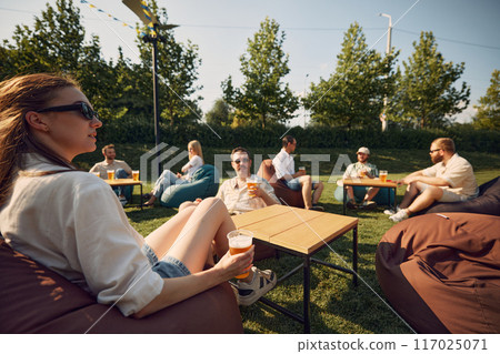 Chilled vibes. Friend, men and women lounging on beanbag chairs, drinking beer, cheerfully talking. Outdoor summer picnic in public place Chilled vibes. Friend, men and women lounging on beanbag chairs, drinking beer, cheerfully talking. Outdoor summer picnic in public place 117025071