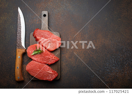 Three raw uncooked meat beef top sirloin steaks on wooden cutting board with rosemary on wooden cutting board with knife on dark rustic background top view, preparing meal with meat, copy space 117025421
