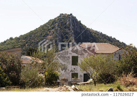 The old building stands surrounded by trees against the backdrop of a big mighty mountain. Southern sunny landscape with an abandoned building. Horizontal 117026027