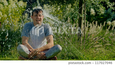 A man is drenched with water while sitting on the grass, looking surprised and amused as he gets soaked on a sunny day in a lush garden. 117027046