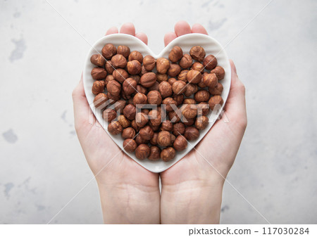 Female hands holding heart shaped plate with peeled hazelnut nuts on light background. 117030284