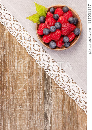 Plate with raspberries and blueberries on the wooden table with laced cloth. 117031137
