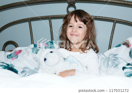 Smiling rested girl is sitting in a bed with sheep toy. 117031272