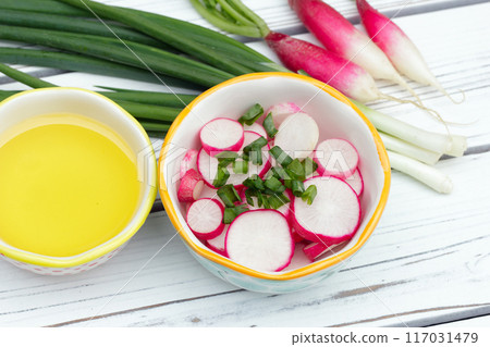 Salad of sliced radishes (French breakfast variety) and green onions in a little bowl, all ingredients with olive oil are on the white wooden rustic table. 117031479