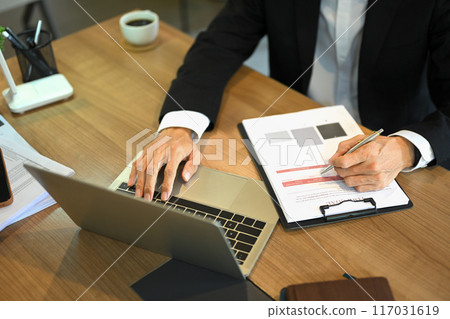 Cropped shot of businessman using laptop and analyzing a finance report at wooden desk 117031619