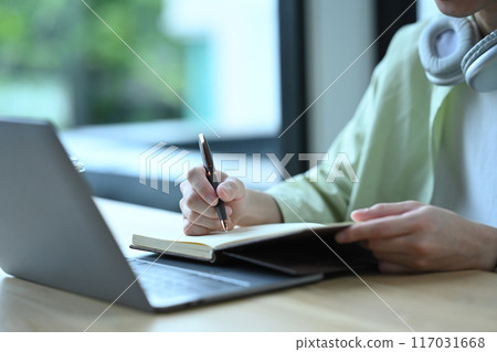 Close up shot of man sitting in front of laptop and handwriting important information on notepad Close up shot of man sitting in front of laptop and handwriting important information on notepad 117031668