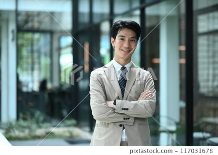 Portrait of smiling businessman standing outside modern office building with arms crossed 117031678