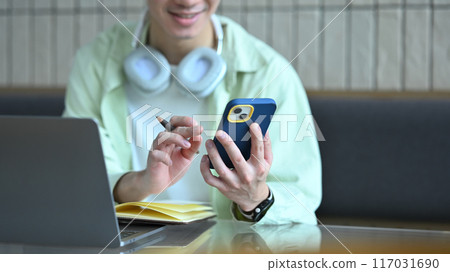 Smiling young man using mobile phone at cafe table, typing message or chatting on social media app Smiling young man using mobile phone at cafe table, typing message or chatting on social media app 117031690