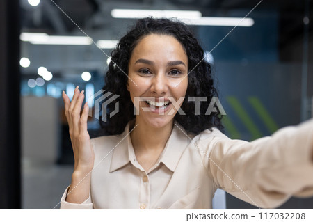 Cheerful businesswoman engaging in video call from office. Confident professional looking at camera with welcoming gesture. Concept of remote work, communication, and modern workplace. 117032208