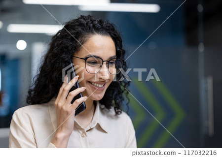 Smiling businesswoman with glasses and curly hair talking on smartphone in modern office. Professional staff engaged in friendly conversation, representing communication and leadership in workplace. Smiling businesswoman with glasses and curly hair talking on smartphone in modern office. Professional staff engaged in friendly conversation, representing communication and leadership in workplace. 117032216