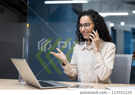 Happy businesswoman with glasses having phone conversation at desk. Smiling professional using laptop and smartphone during work. Engaged in friendly communication in modern office ambiance. Happy businesswoman with glasses having phone conversation at desk. Smiling professional using laptop and smartphone during work. Engaged in friendly communication in modern office ambiance. 117032220