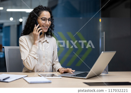 Confident businesswoman smiling during phone call while working on laptop in modern office. Professional office worker multitasking on phone and computer. Workplace communication and productivity 117032221