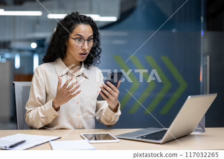 Shocked businesswoman looking at smartphone in office. Woman reacting to surprising or alarming news. Workspace with laptop, tablet, and notepad. Indoor professional setting. 117032265