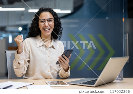Cheerful businesswoman celebrating success in modern office holding smartphone. Happy professional employee expressing achievement and joy while working on laptop. Concept of business victory 117032266