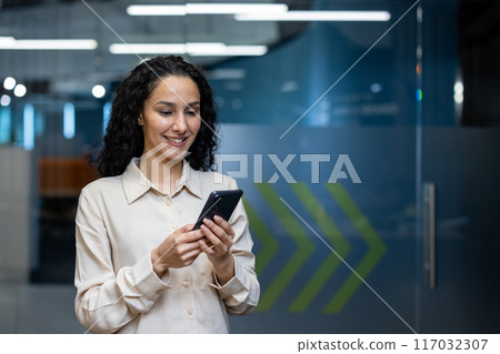Businesswoman standing in modern office using smartphone. Woman smiling and engaging with mobile device in professional workspace, illustrating technology, communication, and professional concepts. 117032307