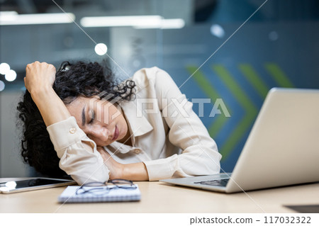 Tired businesswoman resting head on desk in modern office setting. Overwhelmed, stressed out female worker asleep workplace. Concept of workplace fatigue, stress, exhaustion, and professional burnout 117032322