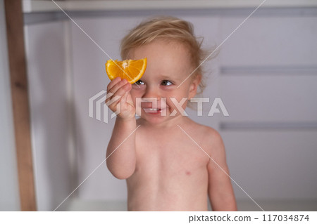 A happy girl 2 years old in the kitchen at home eats delicious sweet oranges. 117034874