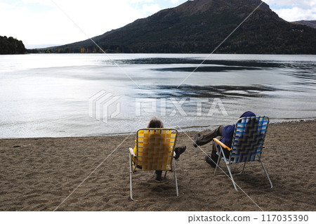 Two people relax on a tranquil lakeside beach, enjoying the scenic mountain view Two people relax on a tranquil lakeside beach, enjoying the scenic mountain view 117035390