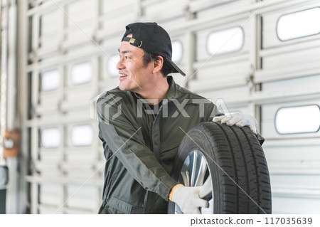一名汽車修理工/機械師的中年亞洲男子拿著輪胎打招呼。 一名汽車修理工/機械師的中年亞洲男子拿著輪胎打招呼。 117035639
