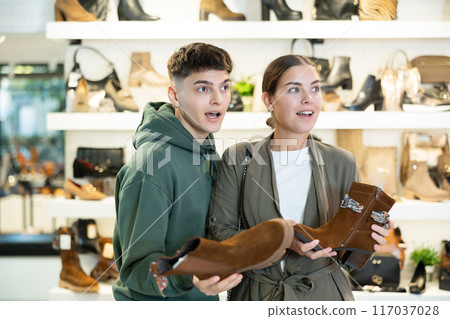 Young guy and girl choosing demi-season womens boots in shoe store Young guy and girl choosing demi-season womens boots in shoe store 117037028