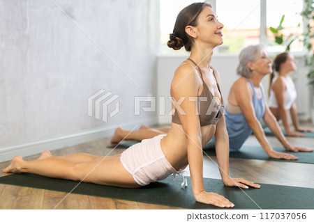 Three generations of women perform upward facing dog pose Urdhva Mukha Svanasana in gym 117037056