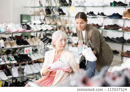 Female sales consultant helps a mature european woman choose shoes sneakers in store 117037324