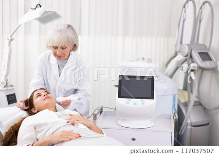 Young female visitor receiving hardware electric lifting massage with help of old female beautician in spa salon indoors 117037507