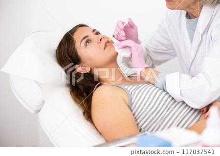 Professional mature female beautician with syringe injecting filler with hyaluronic acid in lips of young woman client during procedure in beauty salon 117037541