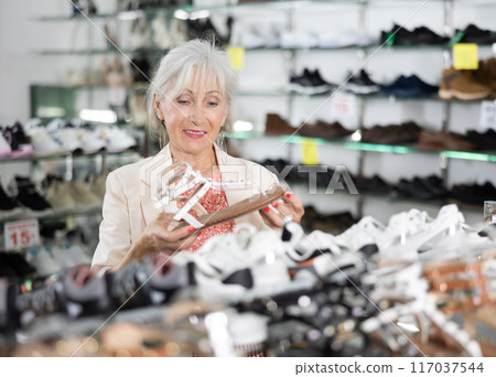 Mature woman client is demanding to inspect sandals in shoe showroom 117037544