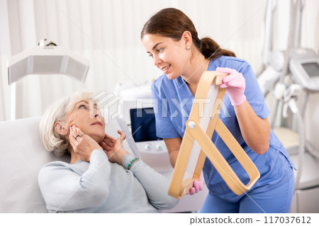 Young female dermatologist showing elderly female client result of facial beauty procedure in mirror in esthetic medicine center 117037612