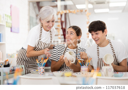 Senior woman assisting happy guy and girl painting ceramic mugs in workshop Senior woman assisting happy guy and girl painting ceramic mugs in workshop 117037618