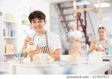 Cheerful skilled young artist painting ceramic cup in pottery workshop Cheerful skilled young artist painting ceramic cup in pottery workshop 117037677