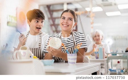 Young couple joyfully talking and painting ceramic mugs in pottery workshop 117037730