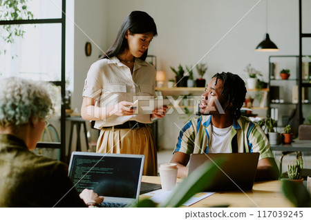 Young Black man and Asian woman wearing casual outfits having conversation while unrecognizable programmer coding on laptop in modern office Young Black man and Asian woman wearing casual outfits having conversation while unrecognizable programmer coding on laptop in modern office 117039245