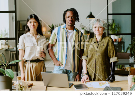 Medium long shot of three diverse gen Z coworkers posing for camera at work in modern eco-friendly office Medium long shot of three diverse gen Z coworkers posing for camera at work in modern eco-friendly office 117039249